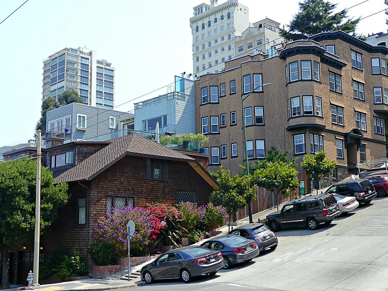 A mixed-use neighborhood with a large apartment building next door to a single-family home on a hilly street with tall buildings looming in the background.