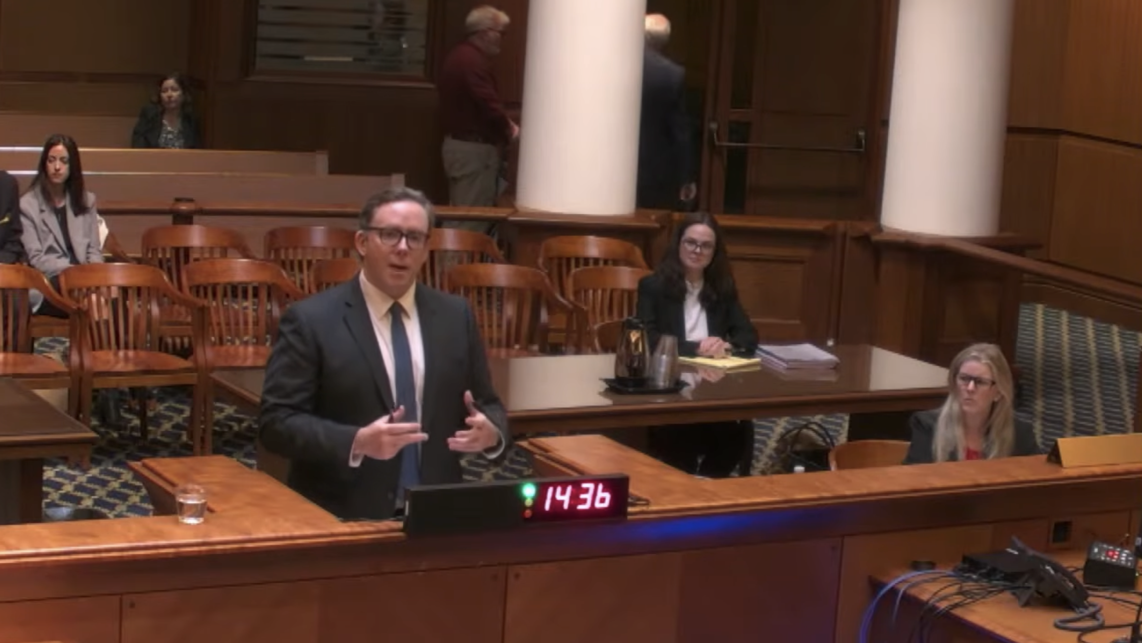 A courtroom scene from the judge’s point of view shows a man in a suit speaking at a lectern. Other attorneys and audience members can be seen in the background.