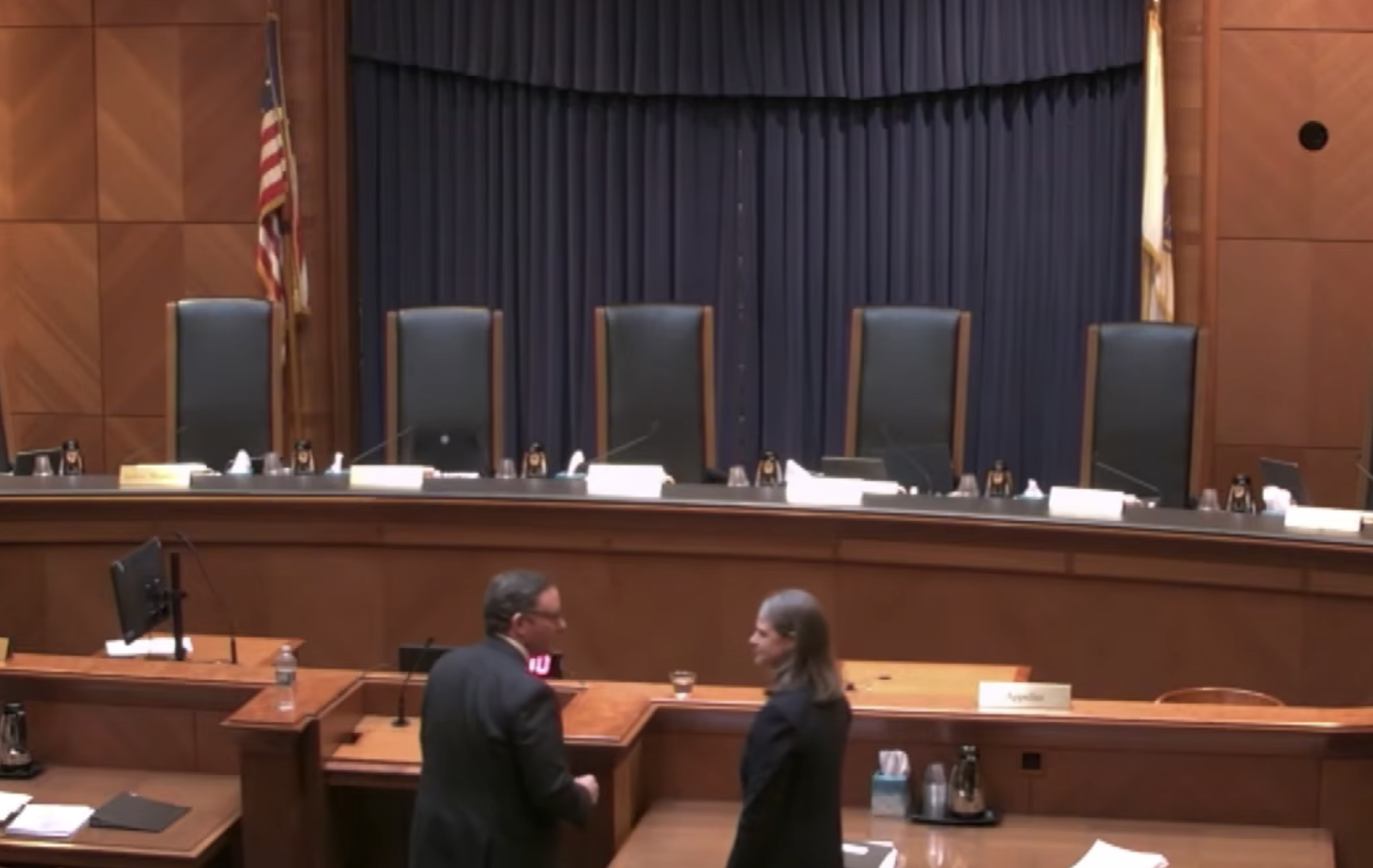 A courtroom scene from the audience’s point of view shows a man in a suit speaking to a woman in a business suit. The justices’ chairs are empty in the background.