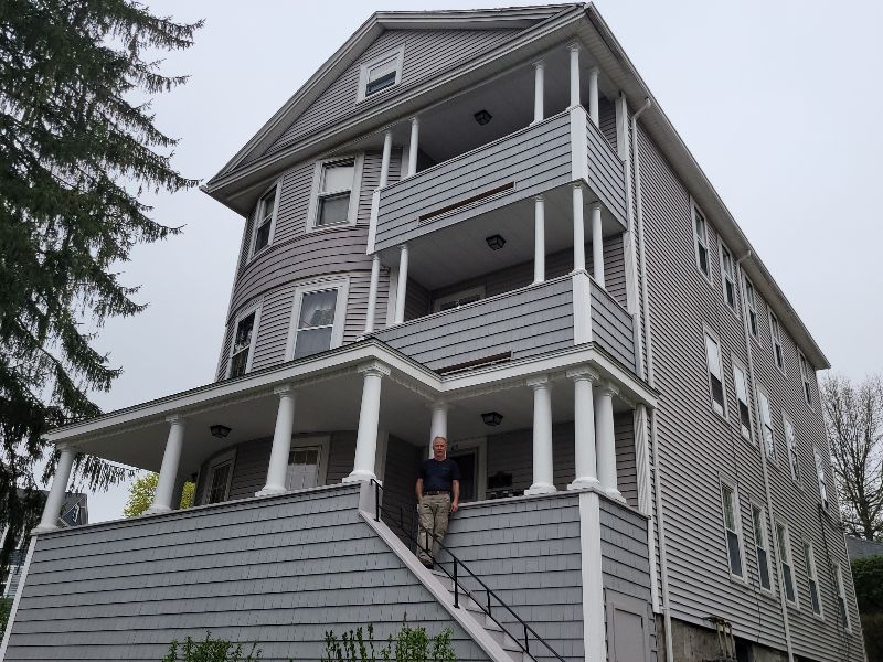 Phil Arsenault is pictured standing on the front stoop of his Worcester triple-decker, a gray-painted multifamily building with white trim.