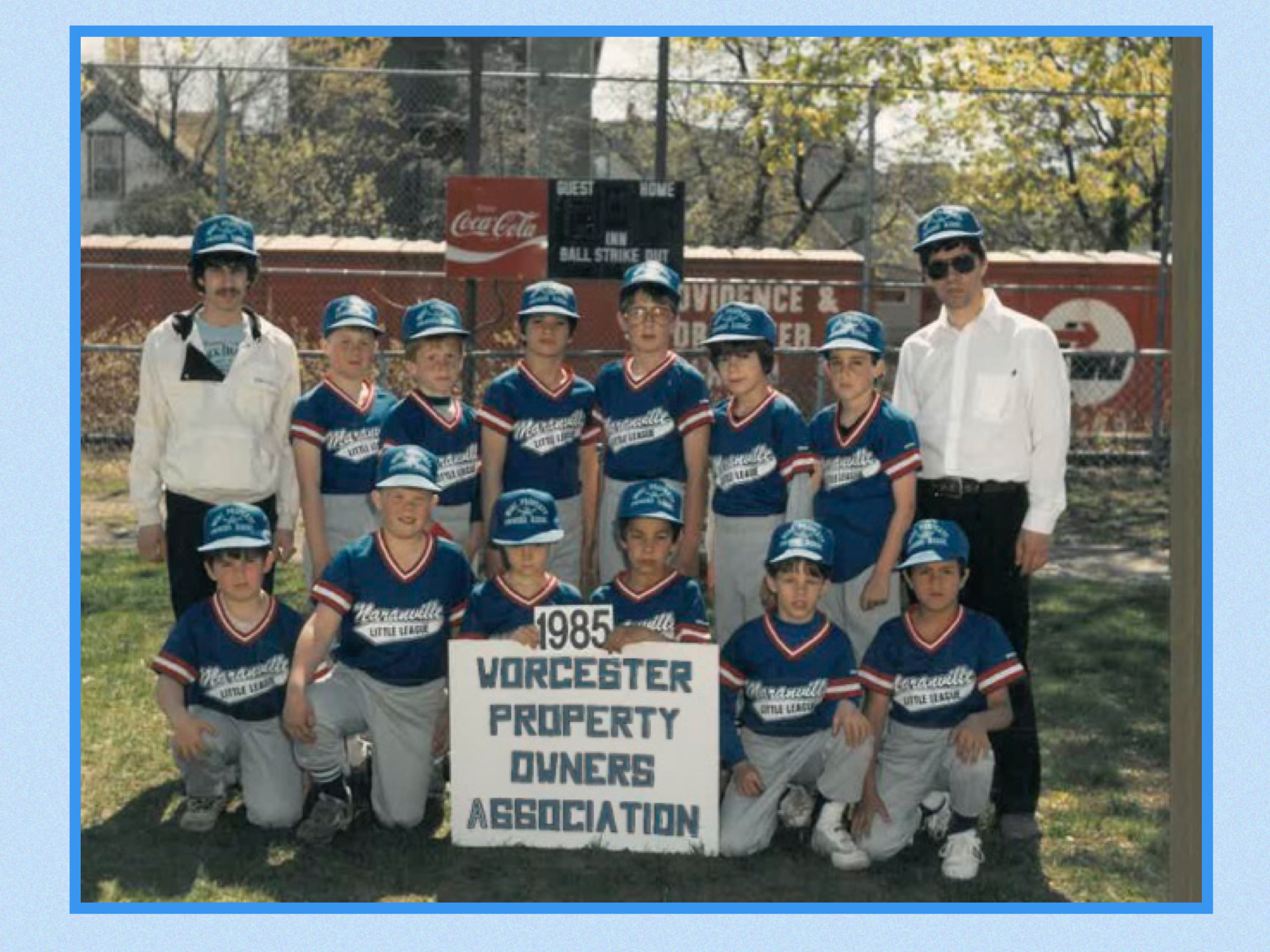 An outdoor photograph from the 1980s featuring a Little League team in blue and gray uniforms. Two adults in blue hats and white shirts/jackets are also in the picture. The sign reads “1985 Worcester Property Owners Association.”