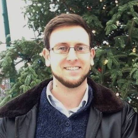 Alec Bewsee smiles in front of a fir tree. He wears a broad collared winter jacket, glasses and a beard.