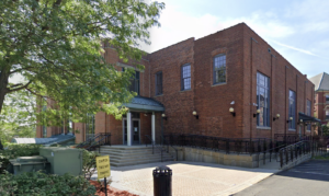 A 2-storey brick building surrounded by trees and a small brick courtyard. A parking lot can be partially seen in the foreground.