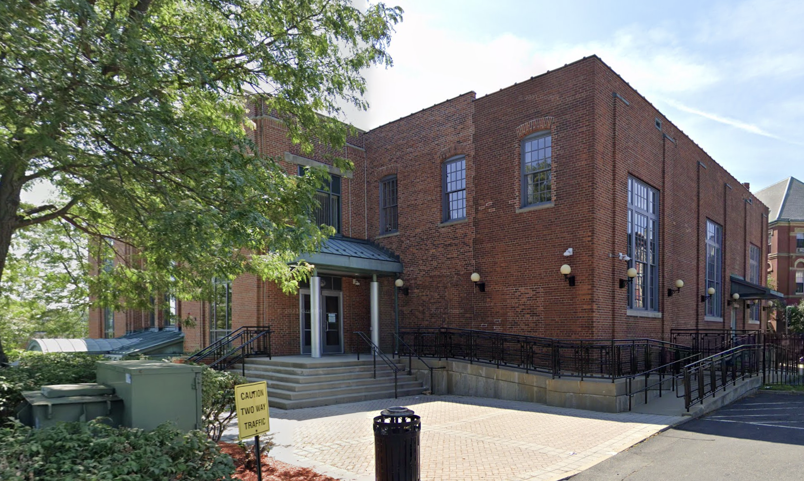 A 2-storey brick building surrounded by trees and a small brick courtyard. A parking lot can be partially seen in the foreground.