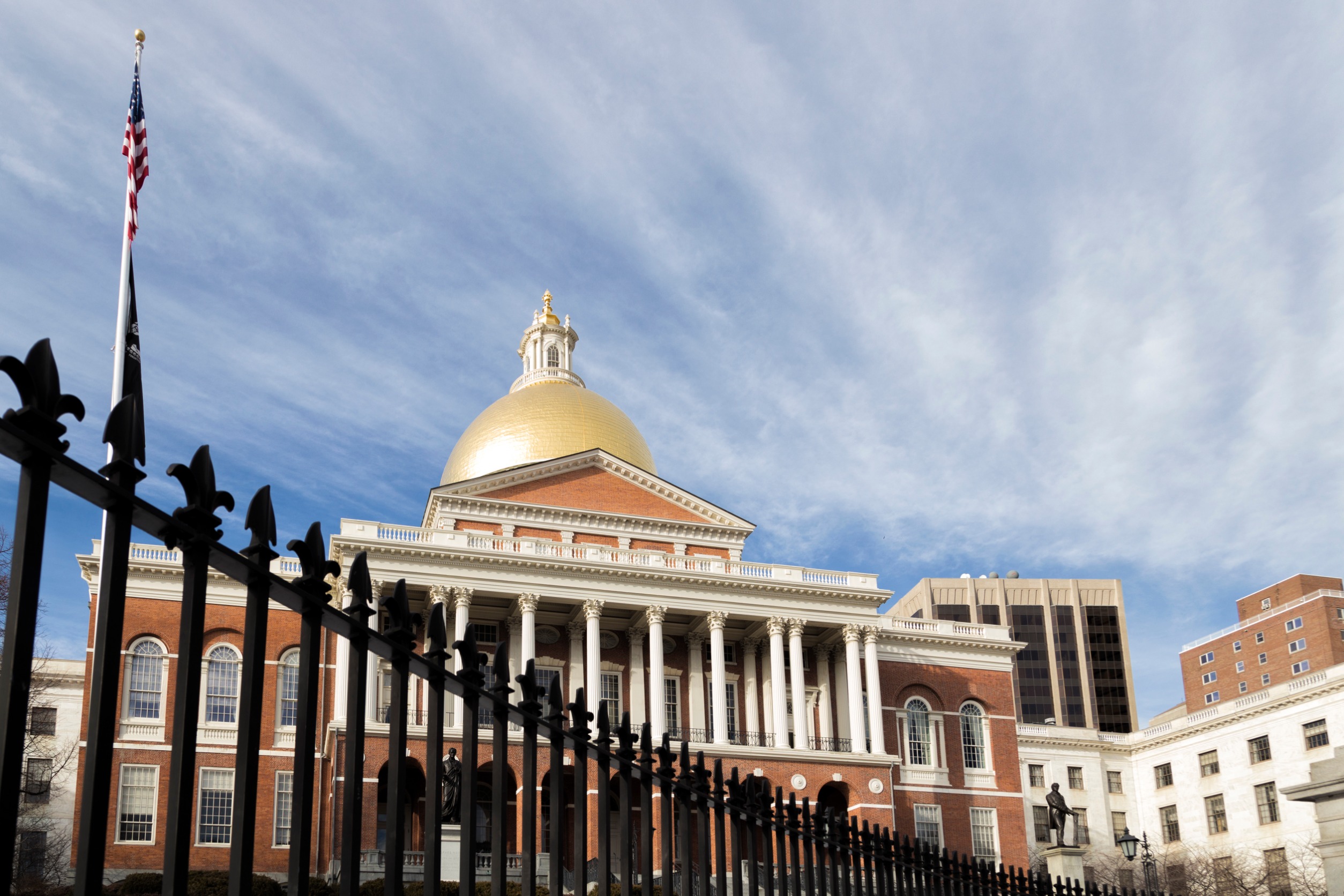 A street view of the Massachusetts State House from outside the gates in winter. It is a brick multi-story building with a gold dome.