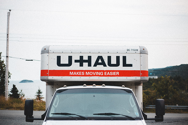 A close up of the front of a U-Haul moving truck driving towards the viewer, with a hazy body of water in the background.