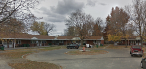 A street view taken in autumn of a row of connected one-story brick apartment buildings at the end of a paved cul-de-sac. Trees with yellow and red leaves surround the buildings.