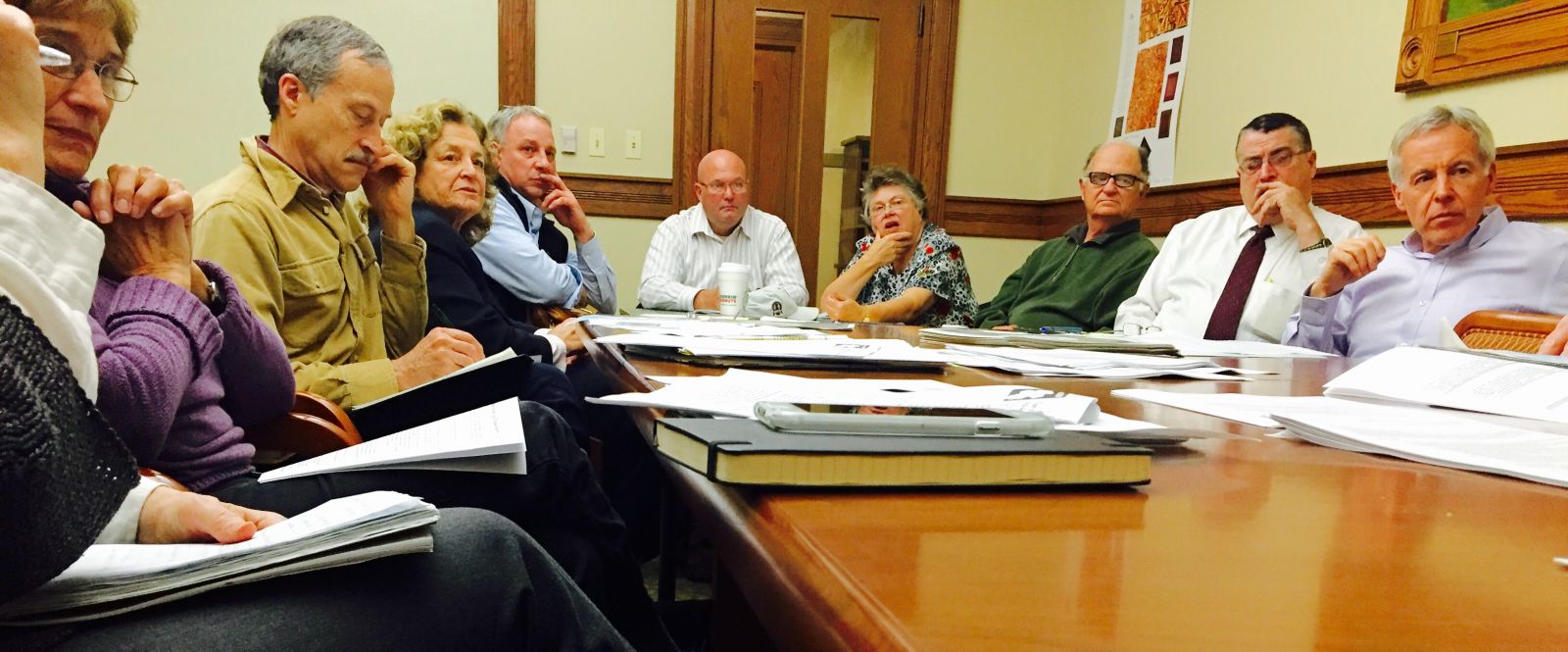 A group of nine people in business casual attire sit around a brown wood rectangular conference table. The person closest to the camera on the left side of the table is not fully in frame. Papers and books are scattered on the table’s surface.]