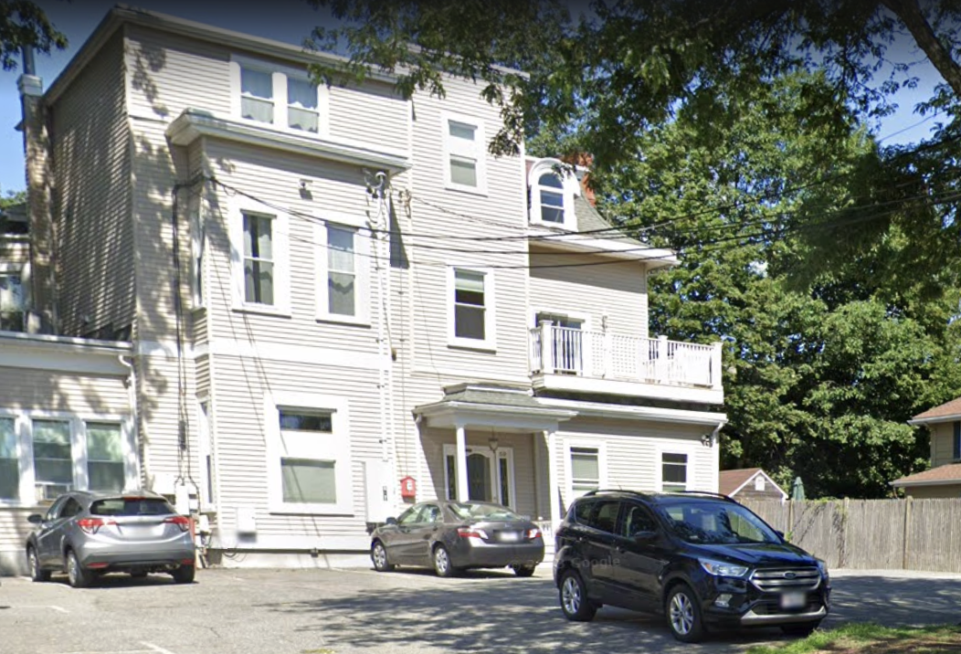 A multi-story multifamily building with beige siding, trees along the driveway and a small deck outside one of the units on the second floor.