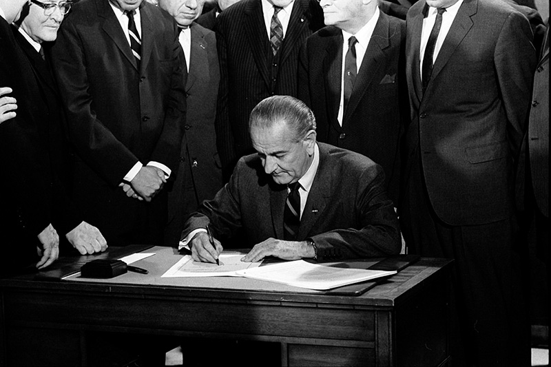 A black and white photo showing President Lyndon B. Johnson sitting at a wooden desk with multiple other white men standing behind him, signing the Fair Housing Act of 1968 into law. 