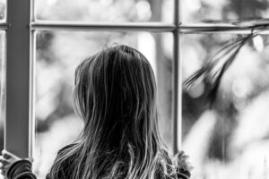 A little girl, photographed in black and white from behind, stands looking out of a window with multiple panes.