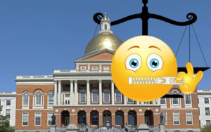 An emoji face holds a finger up its mouth to advise shush as it looms over the State House. The scales of just hang in the background.