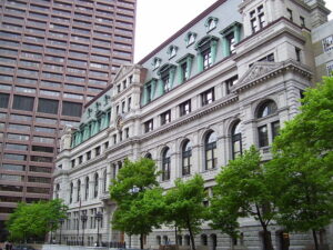 A side view of the John Adams Courthouse, a multistory stone building on a tree-lined sidewalk in Boston.