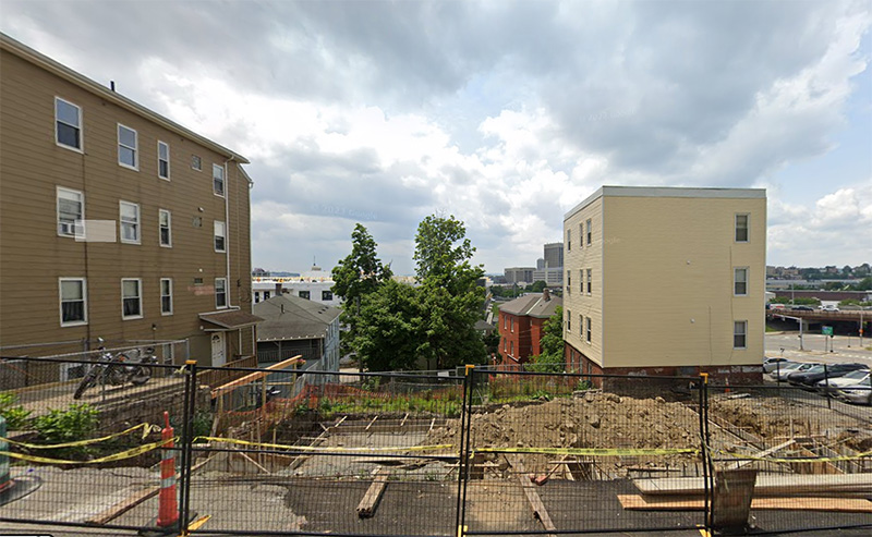 A street view of an empty lot where construction has started: the existing building has been torn down and some wood framing for a new foundation is present. The lot is blocked off with a temporary black construction fence.