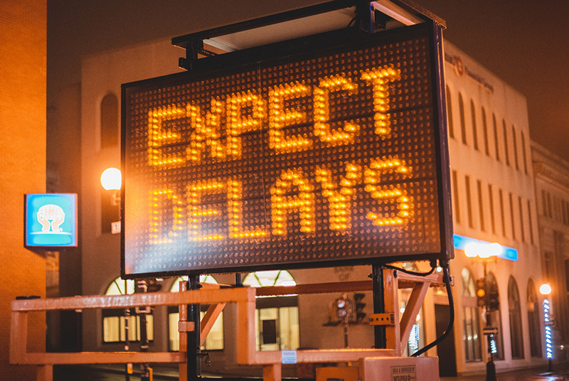 A large digital traffic sign sits on a street in front of buildings in a city. It is nighttime. In big orange letters, the sign reads “Expect Delays.”