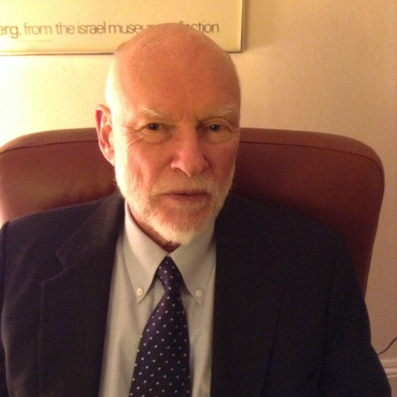 John wears a suit and tie and sits in a leather chair in front of a museum poster.