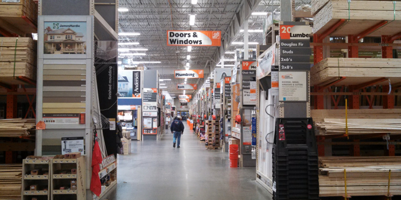 a photo of the interior of a Home Depot store. Shelves of lumber are visible in the foreground, with signs indicating other departments going down the aisle. A person in jeans and a sweatshirt facing away from the camera walks down the main aisle.