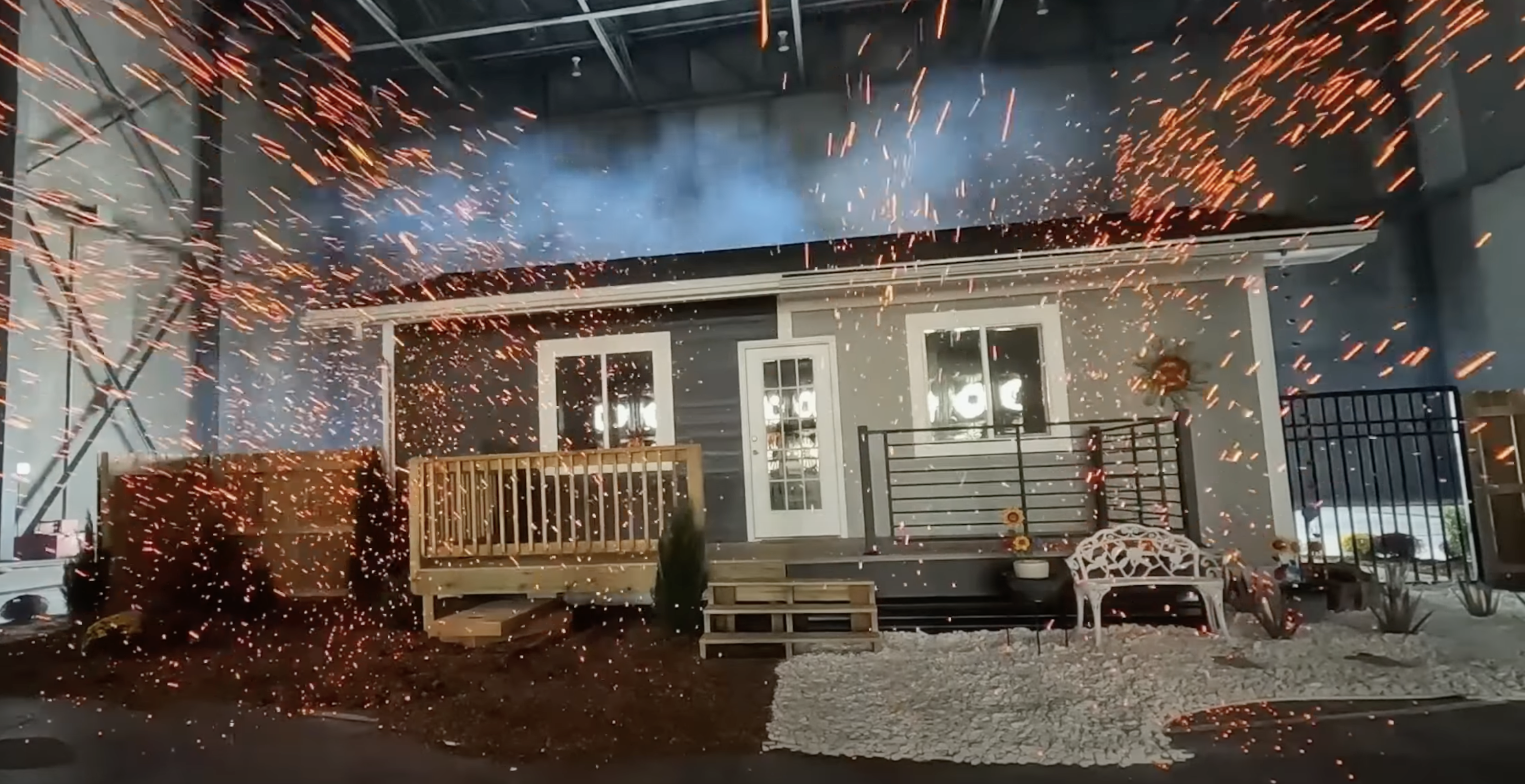 A full-size single story house sits in a warehouse-like room. Embers are flying at the home. The left side of the house has wooden railings and vegetation close to the house. The right side has no close vegetation and metal railings.