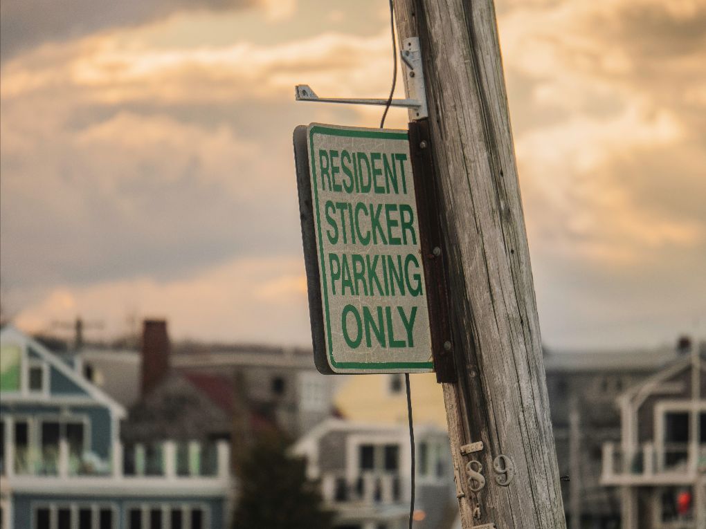 A close-up of a green and white street sign attached to a wooden pole that reads “Resident Sticker Parking Only.” Single-family homes are visible in the background.