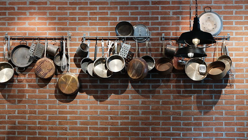 An assortment of pots and pans and related cooking utensils hangs on a black metal rack against a brick wall.