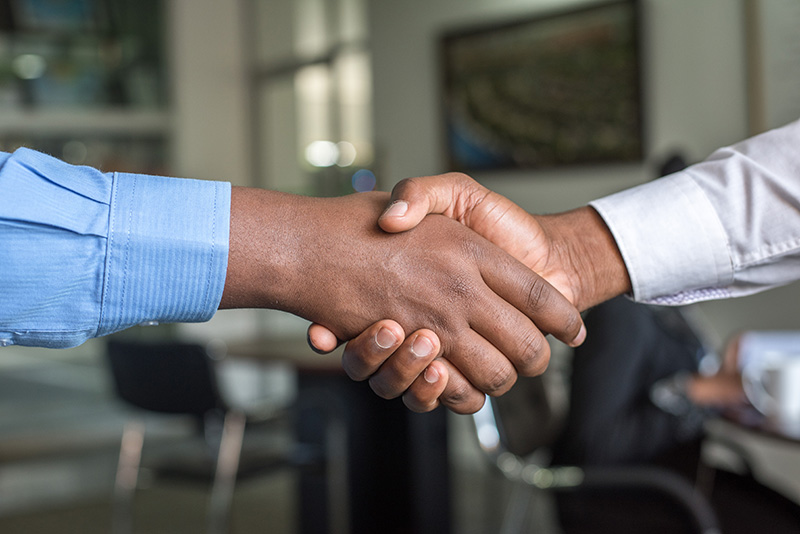 This is a close up of two people in dress shirts shaking hands in an office setting. The person on the left wears a blue dress shirt, the one on the right wears a white dress shirt. Their hands take up most of the image.