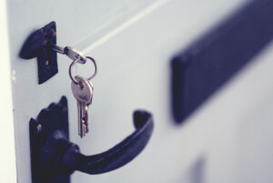 A close-up of a white front door with black lock and handle. A silver key on a ring of other keys hangs from the lock.