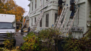 Five ladders lean against a vinyl clad three-decker with four technicians removing individual pieces of siding to drill holes for blow-in cellulose.