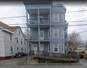 A blue triple-decker home sits close to the street. A wooden accessibility ramp crosses the front of the house at street level. A child’s bicycle is on its side in the small front lawn area.