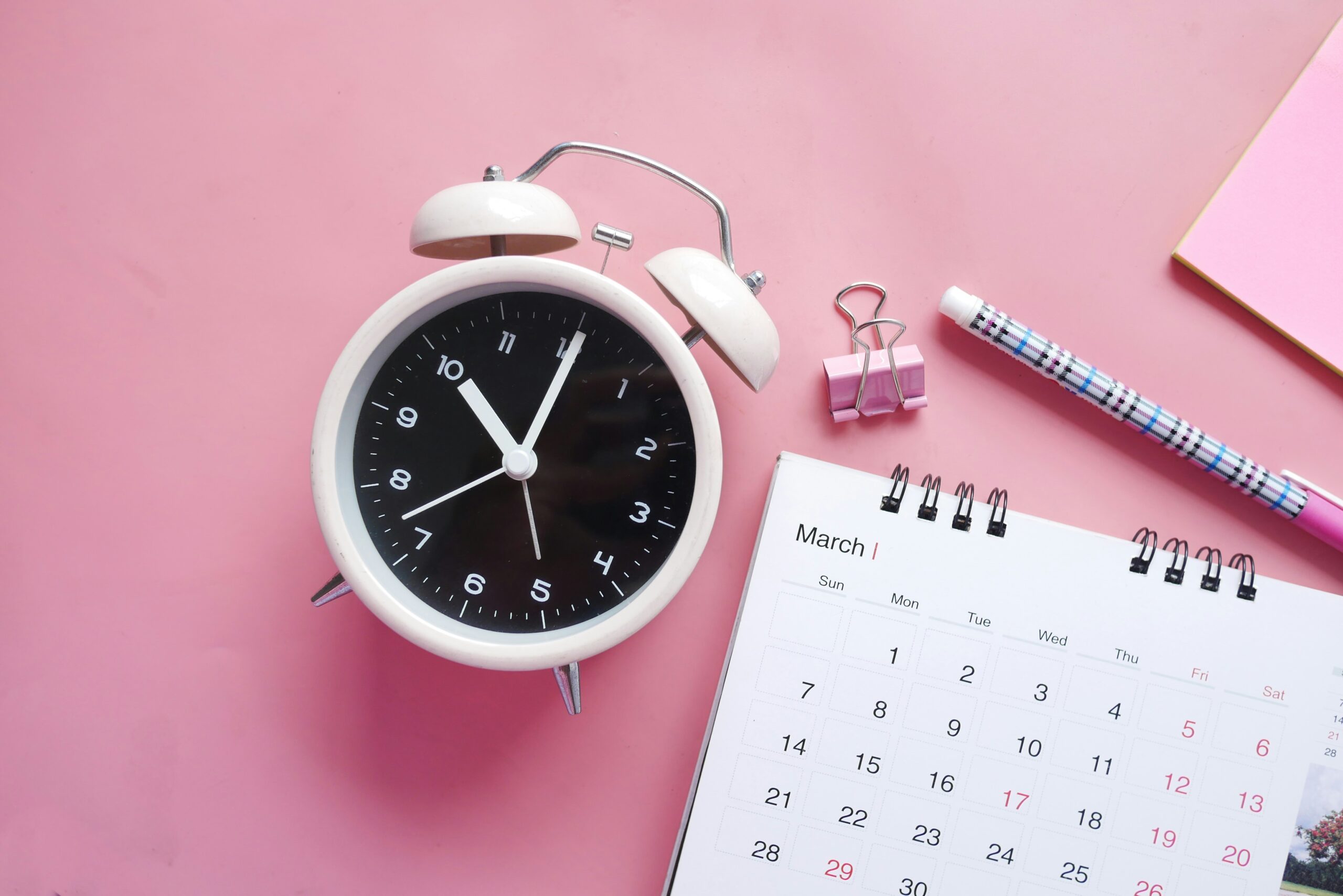 An image of a pale pink alarm clock, a calendar page showing the month of March with no designated year, and a colorful pen, pink binder clip and matching pad of paper against a dark pink background. The clock is set to 10 o’clock.