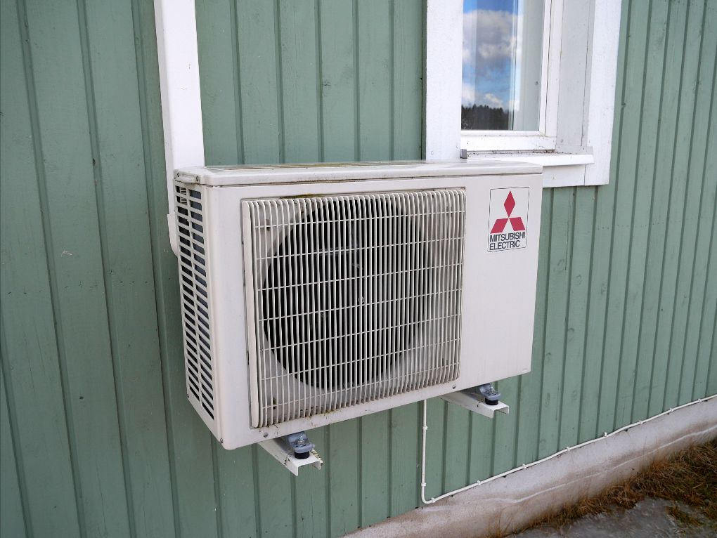 The compressor unit of a Mitsubishi heat pump is mounted against a green paneled exterior home wall.