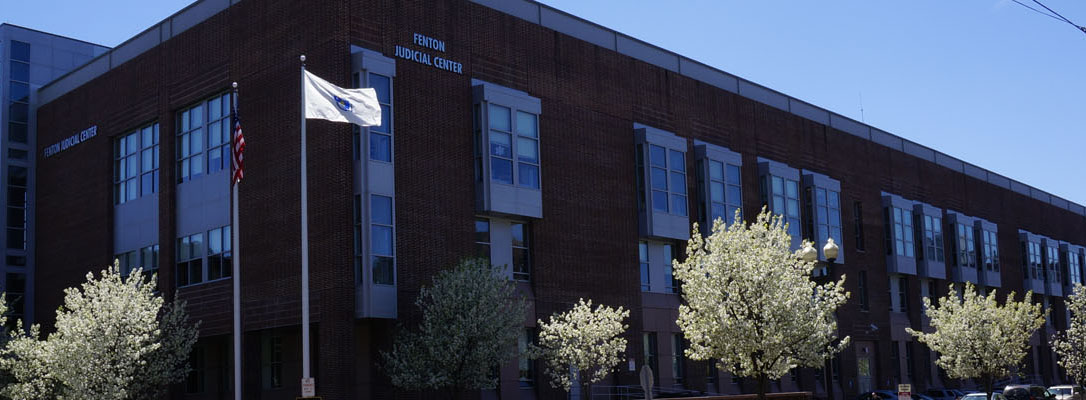 A street view of the Fenton Judicial Center, home of the Lawrence session of the Northeastern Housing Court. It is a multi-story brick building with the American and Massachusetts state flags flying out front.