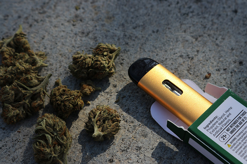 Several nuggets of marijuana sit on a rough concrete surface. To the right is a gold and black oval vape pen half out of its box. The box states the pen contains a solution with “hemp plant extract.”