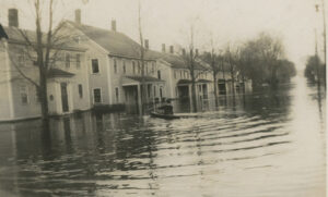 A black and white image of a flooded residential street with water up almost to the front doors of the houses. A man in a rowboat sits with his back to the camera.