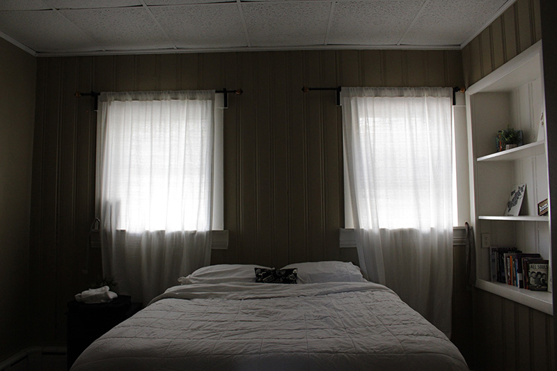 A small bedroom with a dropped ceiling with white tiles. A bed with a white bedspread sits beneath two windows that have sunlight coming in.