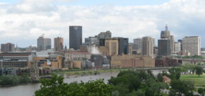 An aerial view photo of the St. Paul, Minnesota, skyline, with multiple buildings and skyscrapers clustered along the Mississippi River in the foreground, with a tree-filled park across the river and a cloud-filled sky in the background.
