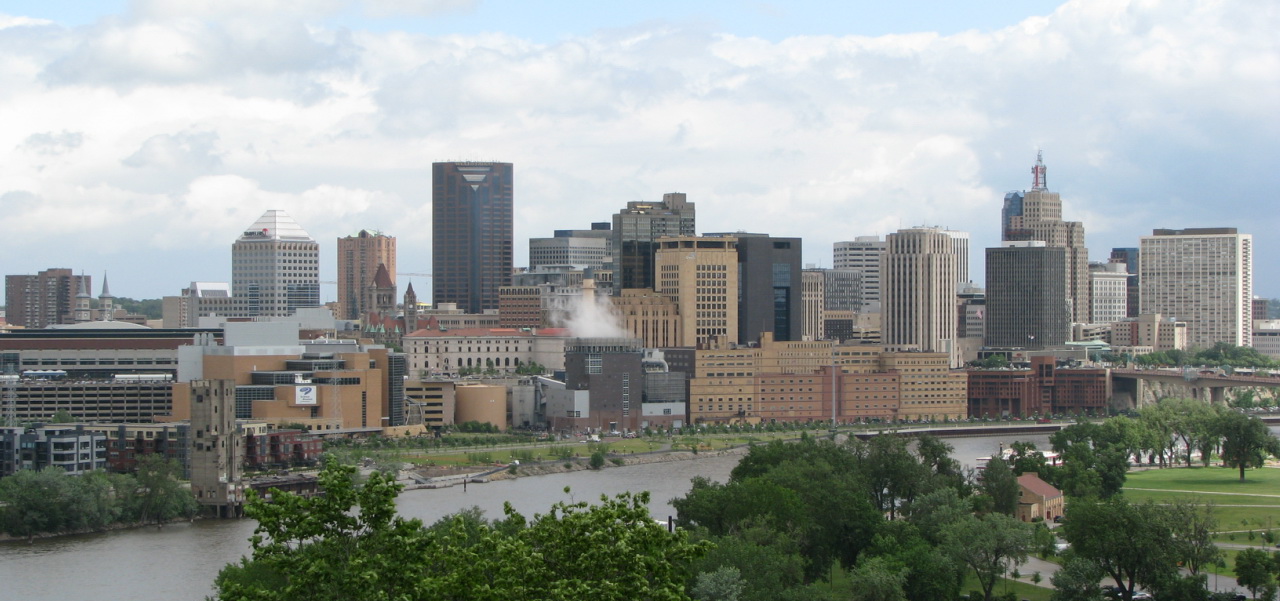 An aerial view photo of the St. Paul, Minnesota, skyline, with multiple buildings and skyscrapers clustered along the Mississippi River in the foreground, with a tree-filled park across the river and a cloud-filled sky in the background.