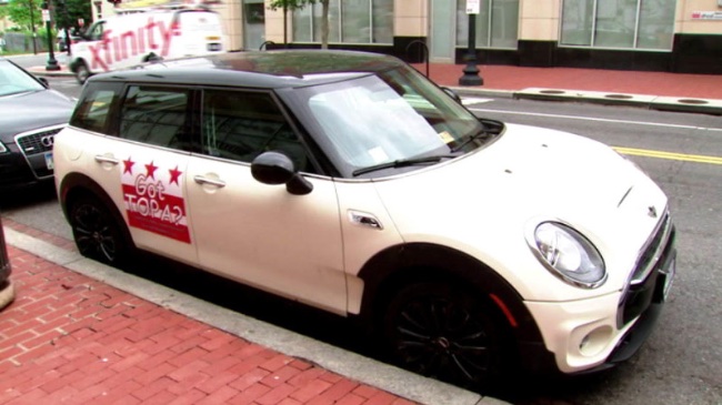 Photograph of a Cooper Mini, white with black top, with a red and white placard stenciled on the back door, reading, “Got TOPA?” parked on a street with brick sidewalk.