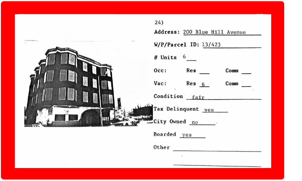 A computer scan of a black and white photo of boarded up, abandoned building, with an identification field to its right, on a red background.