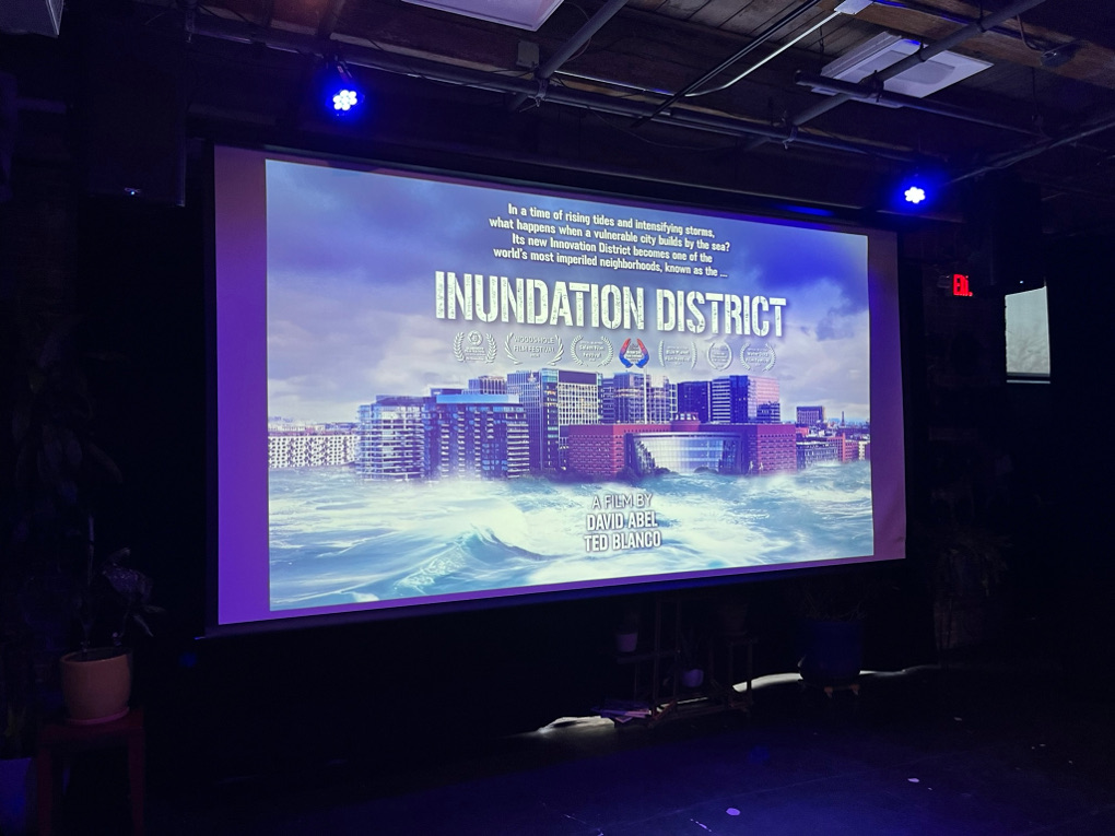A dark room with blue lights frames a screen showing the title screen of Inundation District. The tagline is, âIn a time of rising tides and intensifying storms, what happens when a vulnerable city builds by the sea? Its new Innovation District becomes one of the worldsâ most imperiled neighborhoods, known as the Inundation District.â A film by David Abel, Ted Blanco. The film has been nominated for or won seven awards, the emblems of which are not all legible, including the Woods Hole Film Festival.