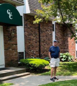 a man with a white moustache, wearing a blue baseball cap, blue t-shirt and white shorts, stands in front of a modern brick apartment building with “CVC” stenciled on an awning, manicured lawn and trimmed bushes, on a sunny day.