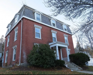 A street view of a two-story brick house with a grey roof, three attic windows and a round classical-style white porch. There are tall shrubs in front of the house and tree branches are visible in the upper right corner of the image.