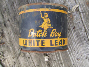 A photo of a Dutch Boy White Lead paint can on a wooden floor, with the long-time emblem of the brand, a “dutch boy” sitting on a plank holding a paint can and paint brush.