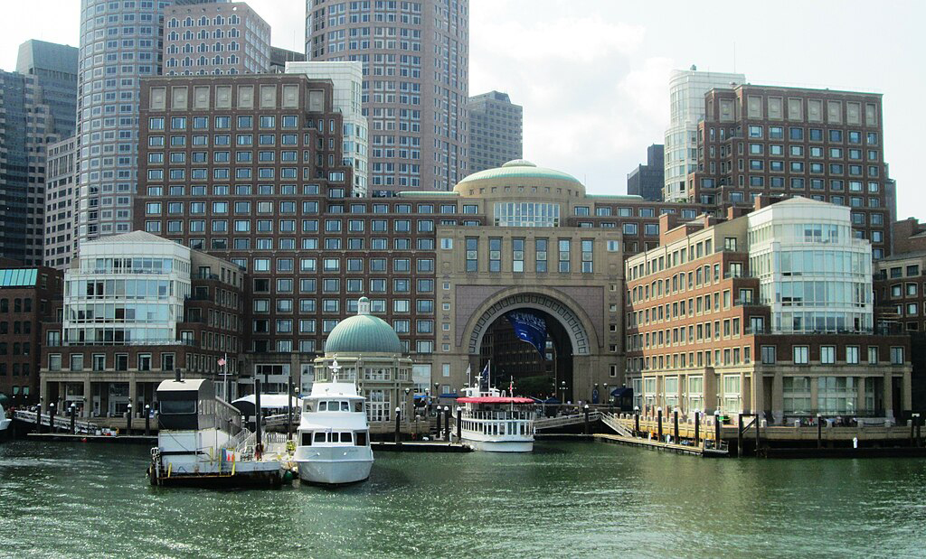 photo of Boston Harbor, with yachts and tourist boats in the water, and towering buildings of downtown Boston in the background.