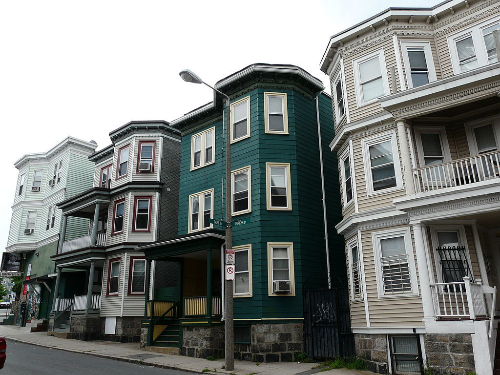 Photo of a row of four triple-decker multifamily homes, painted shades of green and tan, on a Boston street on a cloudy day.