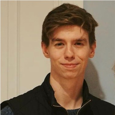 Ryan Bradley smiles at the camera. He is inside a home with wood trim. He wears a sweater vest with high collar and zippered front, is clean-shaven and has wavy hair.