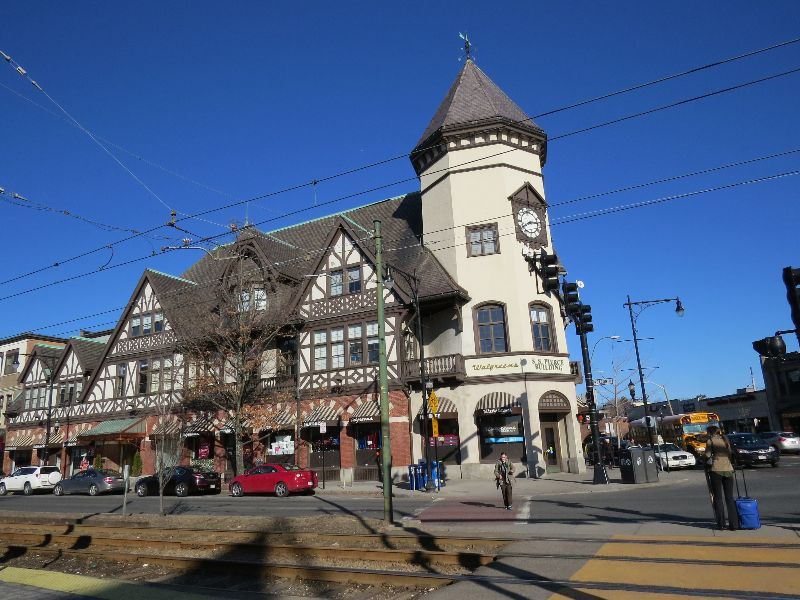 photo of Coolidge Corner, a thriving commercial intersection in Brookline, picturing a block with tudor castle-like architecture and ground-floor shops like Walgreens and others, pedestrians crossing the street and several cars parked along the curb.