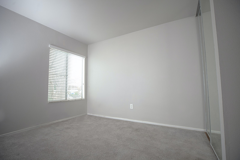 This photograph shows an empty room with a beige carpet and white walls and ceiling. One wall has a window with blinds that are partially open, the wall opposite it appears to have a mirrored closet door.