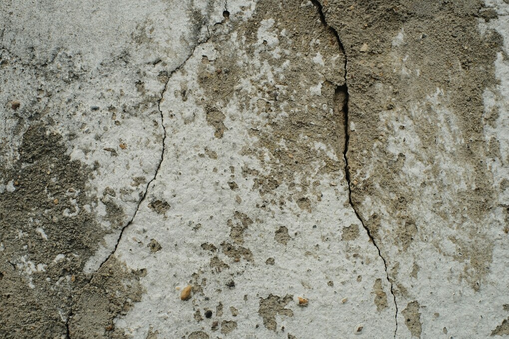 A close-up photograph of a concrete wall with a spiderweb network of cracks, some of which go deep into the stone.