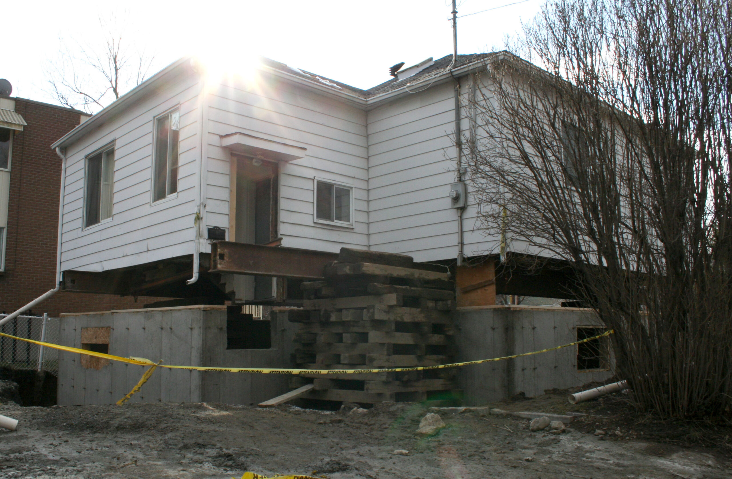 A photograph of a small one-story house with white siding raised up above what appears to be a newly-poured foundation.