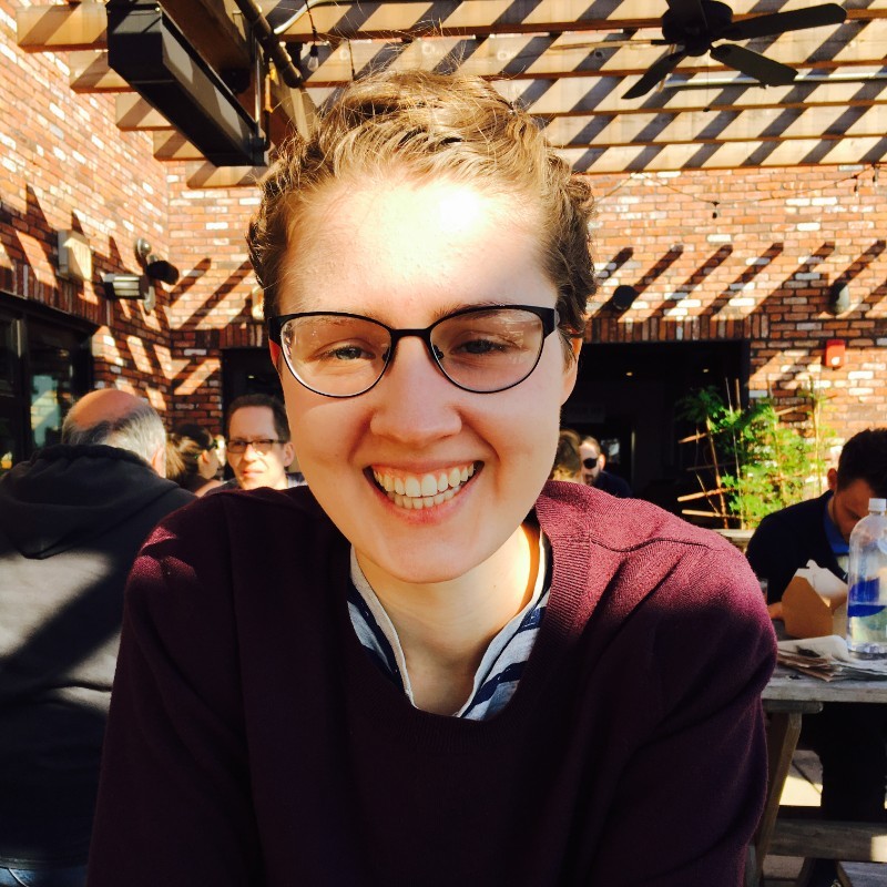 Jenna wears her hair back, wears glasses and smiles broadly in an outdoor café under a pergola.